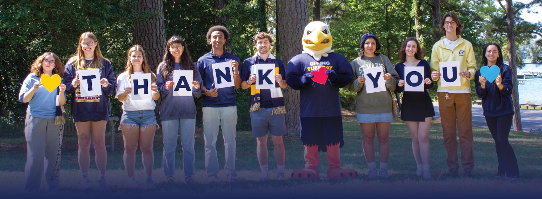 A group of people and a mascot stand outdoors holding signs that spell "Thank You," with heart shapes on either side.