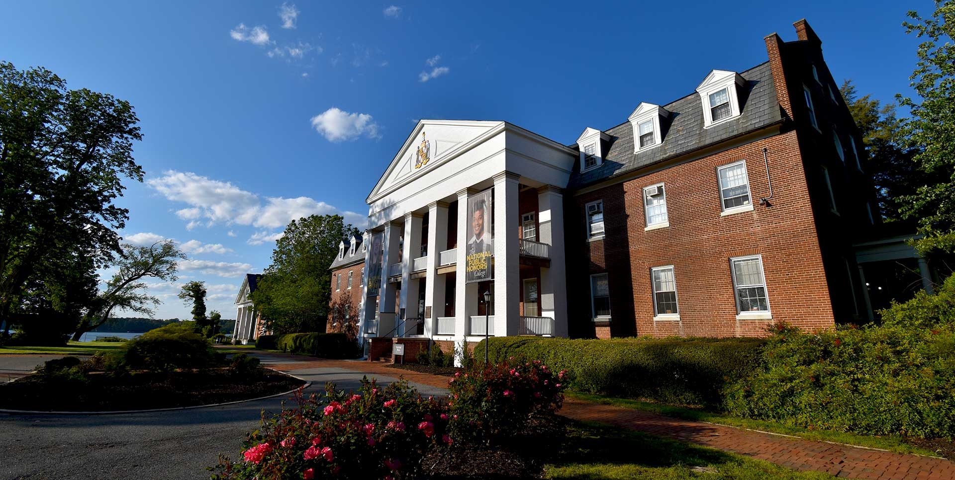 A large brick building with white columns stands under a clear blue sky. The structure has a traditional design with a gabled roof and dormer windows. In the foreground, there's a circular driveway and well-maintained gardens with blooming roses.