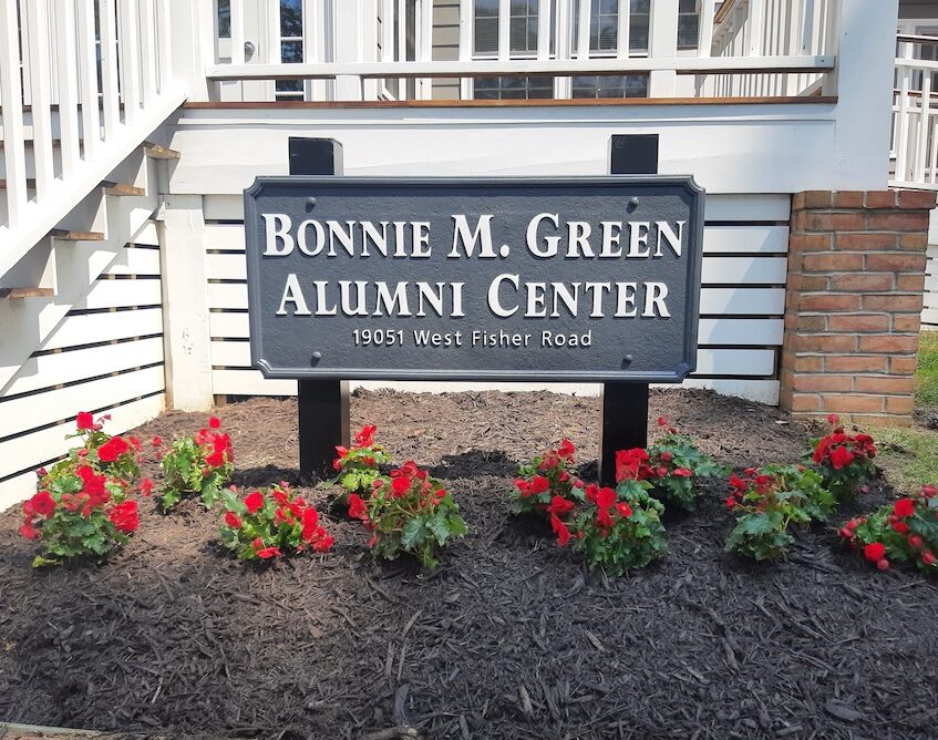 A sign reading "Bonnie M. Green Alumni Center, 19051 West Fisher Road" stands in front of red flowers and mulch by a building with white railings and brick columns.