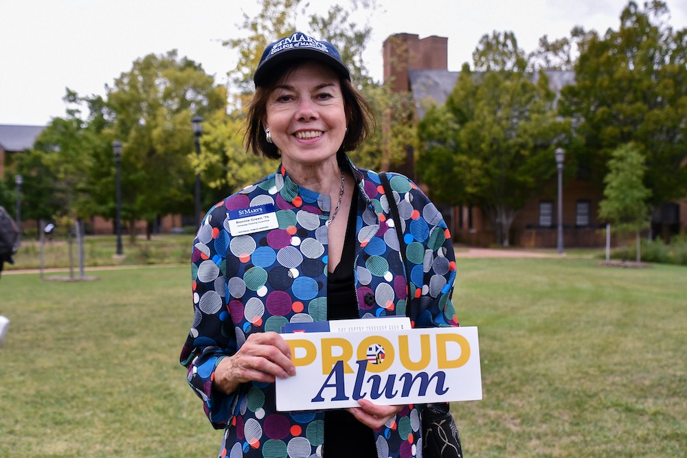 A woman wearing a patterned jacket and name badge stands outdoors, holding a sign that says "Proud Alum." Trees and buildings are in the background.