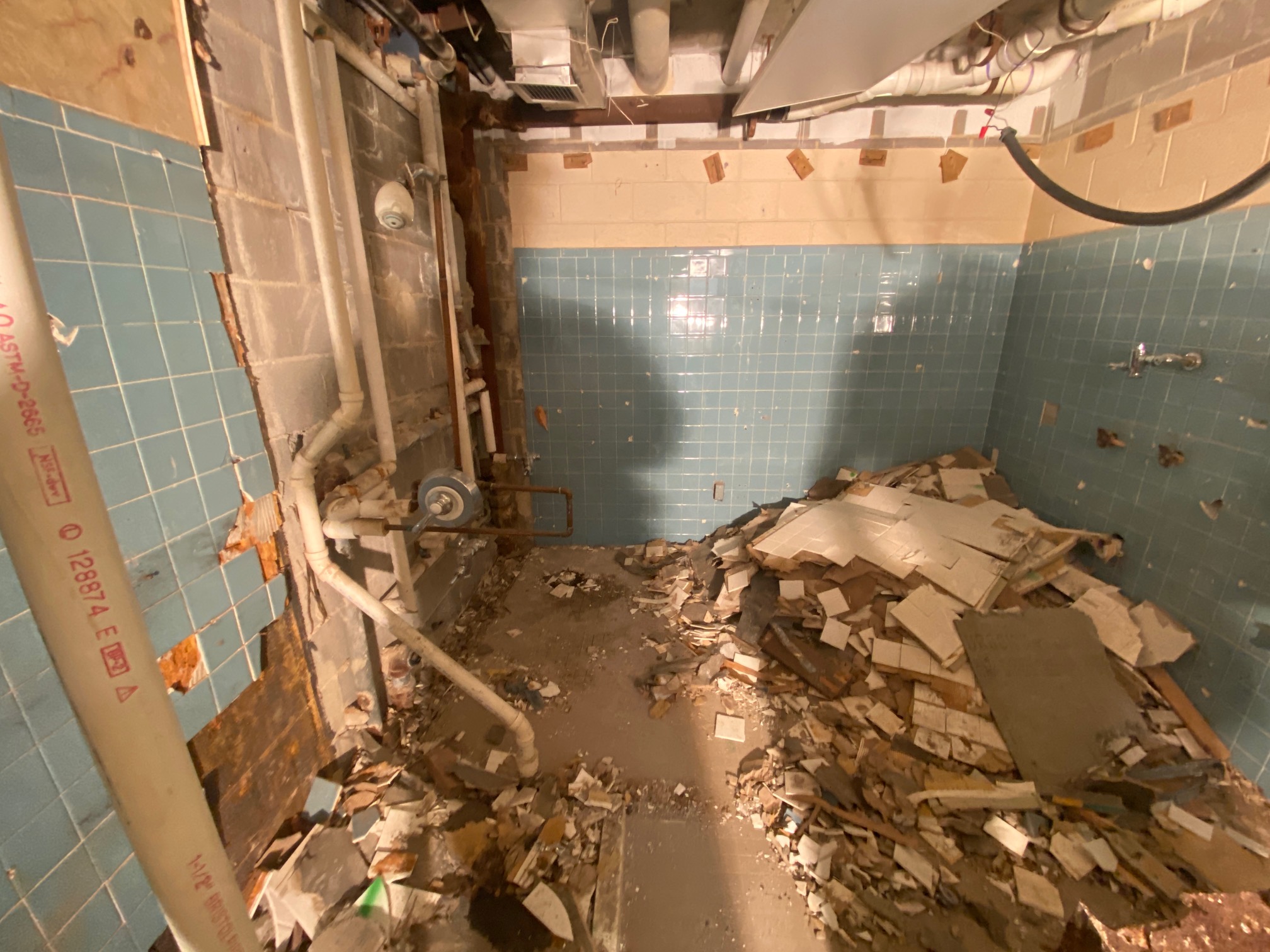 A room with blue tiled walls, exposed pipes, and debris scattered on the floor. The wall tiles and plaster are broken and partially removed, revealing underlying cinder blocks.