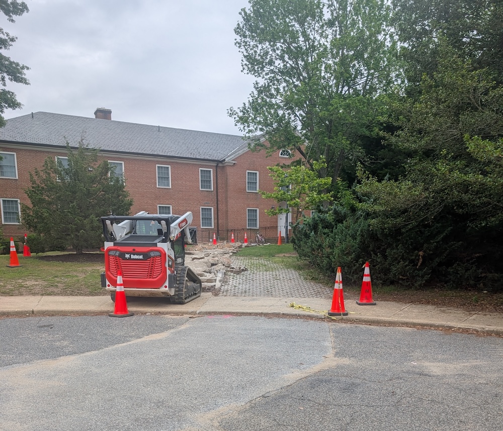 PXL_20250512_183134470.NIGHT A small construction vehicle and orange cones surround a torn-up walkway in front of a brick building with trees nearby.